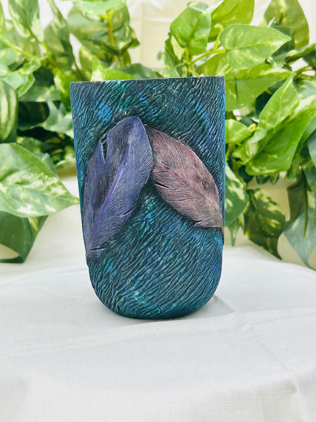 Teal textured vase with feathers on a white surface with green leaves in the background
